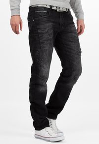 Jeans in denim nero con dettagli strappati, fit tapered e cinque tasche. Indossati con sneakers bianche e un maglione grigio chiaro.