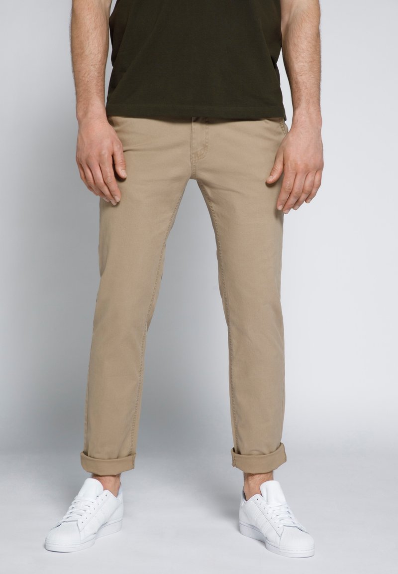 Chino beige con orli arrotolati, realizzati in un morbido misto cotone, abbinati a sneakers bianche. Il design presenta tasche standard e passanti per la cintura.