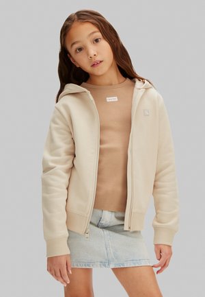 Fată tânără purtând o hanorac bej cu fermoar, un tricou Calvin Klein de culoare bronz și o fustă din denim albastru deschis, stând pe un fundal gri simplu.