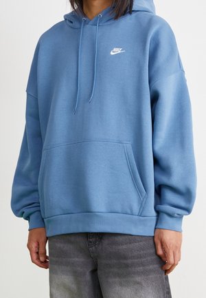 Person iført en blå Nike-hættetrøje med lomme foran og snore, kombineret med falmede sorte jeans.