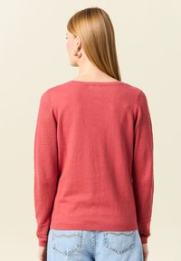 Pull rouge à manches longues avec un corps uni et des panneaux en maille texturée sur les manches. Encolure ras du cou et poignets côtelés. Tissu léger.