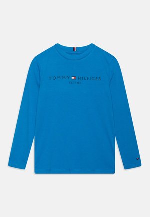 Camisa azul de manga larga con el texto "Tommy Hilfiger EST. 1985" y logo de bandera en el pecho, y pequeño logo de bandera en el puño de la manga izquierda.
