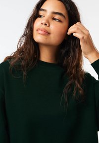 Tmavozelená mikina vyrobená z mäkkej látky, s okrúhlym výstrihom a voľným strihom. Vlasy sú vlnité a po plecia. Prírodný make-up.