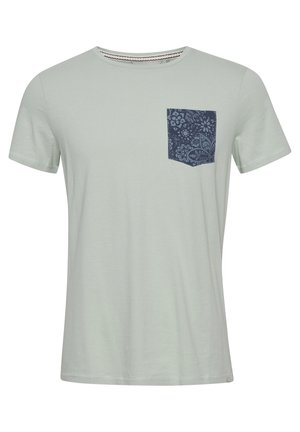 T-shirt vert clair avec un col rond, des manches courtes et une poche à motif bleu marine sur le côté gauche. Texture douce et lisse.