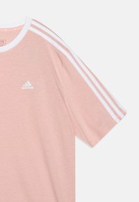 Camiseta Adidas de color rosa claro con detalles de tres rayas blancas en los hombros y un pequeño logo blanco en el pecho. Tela suave y casual.