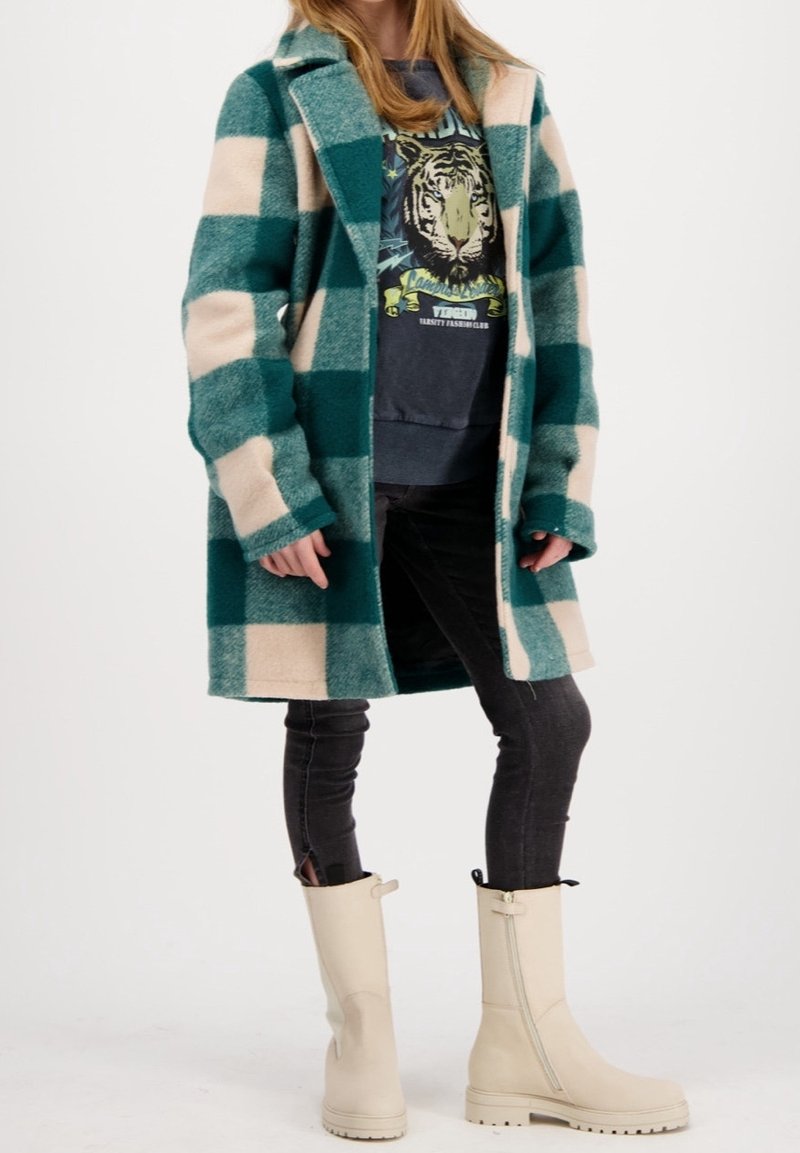 Cappotto di lana a quadri verde e beige con maniche lunghe, abbinato a un hoodie grigio con grafica e jeans neri. Stivali color crema fino al ginocchio.