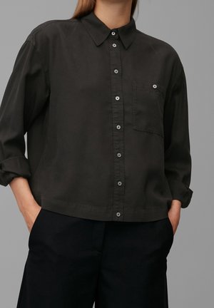 Osoba ubrana w czarną koszulę zapinaną na guziki z kieszenią na piersi i podwiniętymi rękawami, z rękami w kieszeniach czarnych spodni.