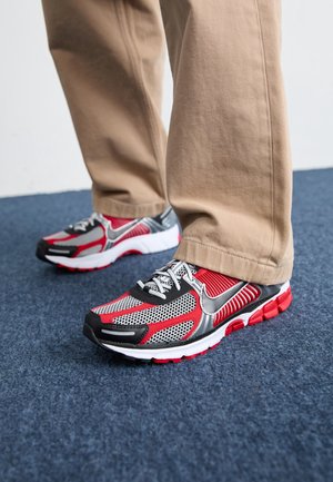 Osoba stoi na niebieskim dywanie, nosząc beżowe spodnie oraz czerwone, czarne i białe buty do biegania Nike z białymi sznurowadłami.