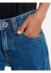Blaue Denim-Jeans mit hoher Taille, ausgestattet mit Kupferbeschlägen, orangefarbenen Nähten und einer Vordertasche. Der Stoff hat eine leicht ausgewaschene Textur.