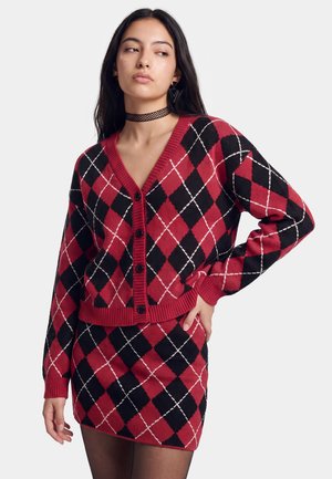 Vrouw draagt een rood en zwart argyle vest en bijpassende rok met zwarte panty's, staand met één hand op haar heup.