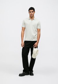 Polo grigio chiaro, pantaloni neri, che tiene un cappellino bianco. Maniche corte, senza motivi visibili, design semplice e vestibilità comoda. Scarpe nere.
