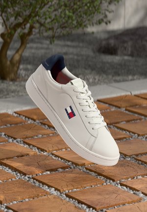 Weißer Sneaker mit niedrigem Schaft, navyfarbener Fersenkappe und Tommy Hilfiger Flaggenlogo, präsentiert auf Holzfliesen und kleinen grauen Steinen im Freien.