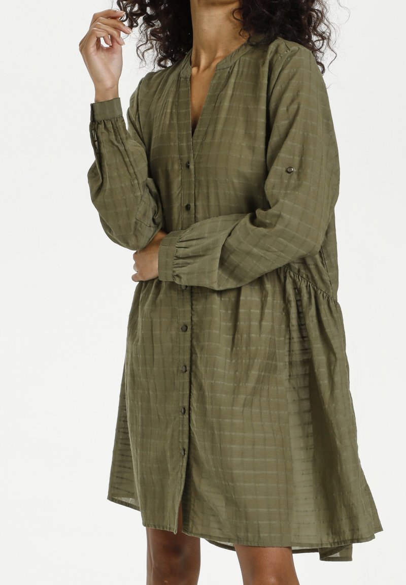 Femme portant une robe longue à manches longues boutonnée, ample, de couleur vert olive avec un motif à carreaux subtil, debout avec un bras levé près du visage.