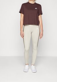 Brun kortärmad t-shirt med vita ränder på ärmarna och logotyp, i kombination med ljusbeiga leggings med sidodetaljer och vita sneakers.