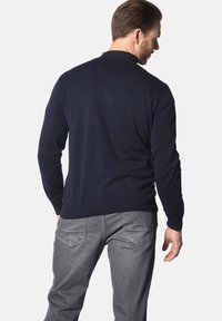 Marineblaues Strickpullover mit geripptem Kragen und Bündchen, kombiniert mit hellgrauen Jeans. Der Stoff wirkt glatt und körperbetont.