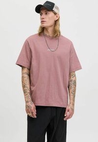 Lyserød t-shirt med løs pasform, korte ærmer og et lille logo i midten. Bæres med sorte bukser og en sort og beige kasket.