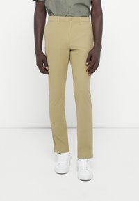 Pantalones beige ajustados con una textura suave, corte slim y diseño de frente plano, combinados con zapatillas blancas y una camisa verde con estampado.
