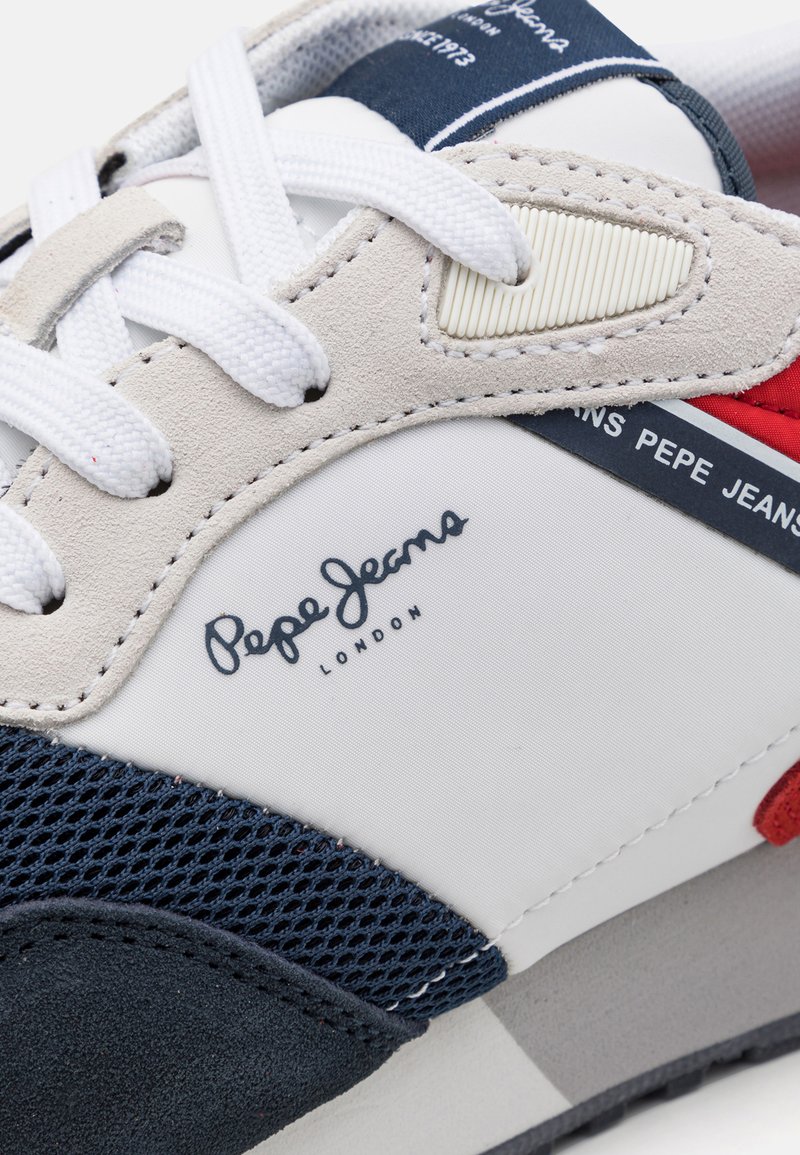 Sneaker mit einer Kombination aus verschiedenen Materialien, darunter Mesh, Wildleder und Synthetik. Die Farben umfassen Weiß, Marineblau und rote Akzente, mit einem markiertem Logo-Detail.