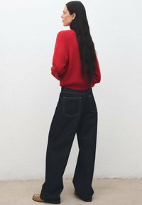 Donna con lunghi capelli neri che indossa un maglione rosso, jeans larghi scuri e scarpe marroni, in piedi contro un muro bianco e rivolta verso sinistra.