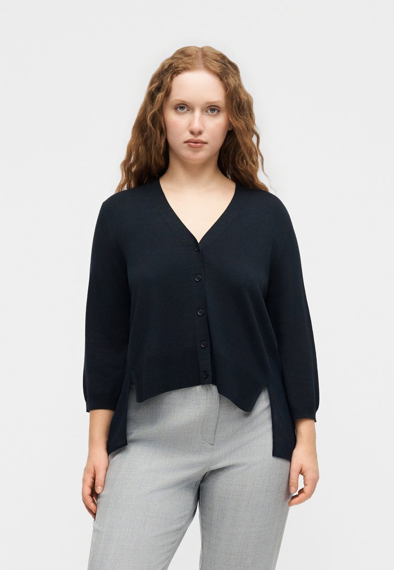 Γυναίκα με μακριά σγουρά μαλλιά, που φοράει μαύρη ζακέτα με κουμπιά και V-λαιμόκοψη και ανοιχτό γκρι παντελόνι, στέκεται μπροστά σε απλό λευκό φόντο.