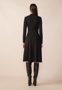 Černé dlouhé šaty s rozšířenou sukní a přiléhavým pasem, v kombinaci s vysokými černými koženými botami. Jednoduchá tkanina, minimální design.