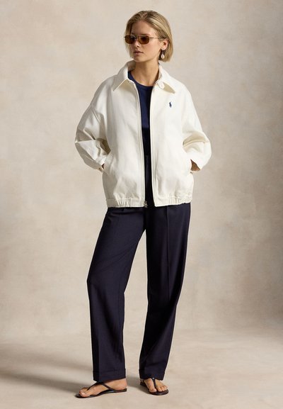 Γυναίκα με γυαλιά ηλίου, λευκό φούτερ με φερμουάρ, μπλε μπλούζα και παντελόνι, στέκεται με τα χέρια στις τσέπες και φοράει μαύρα σανδάλια.