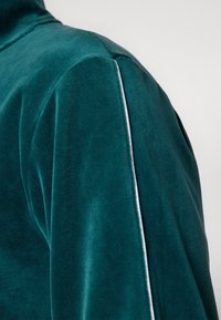 Zöldeskék bársony blúz magas gallérral és fehér díszcsíkkal a vállvarrás mentén, amely puha textúrát és gazdag színt mutat.