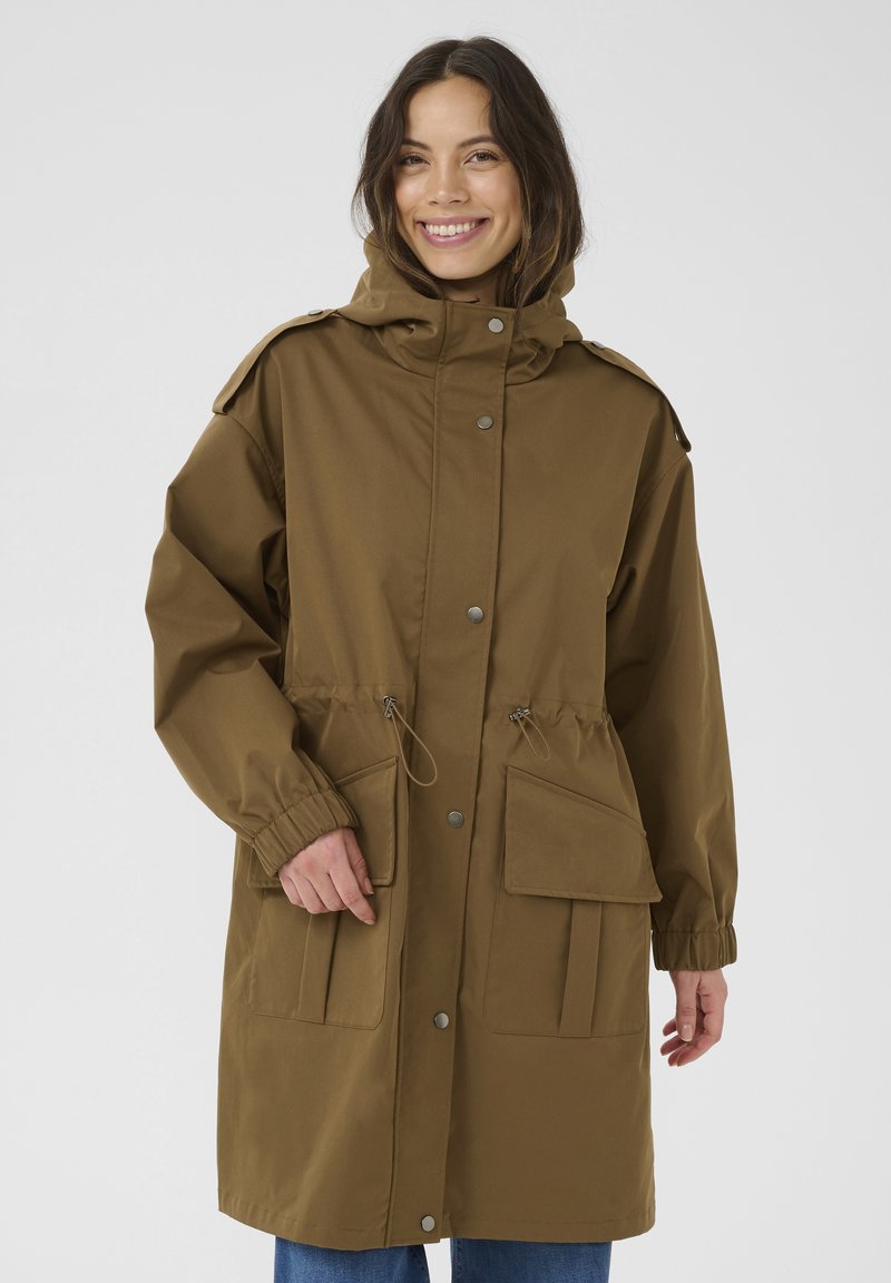 Bruine, lichte parka met een capuchon, trekkoord in de taille, drukknoopjes en twee voorzakken; heeft elastische manchetten en schouderepauletten.