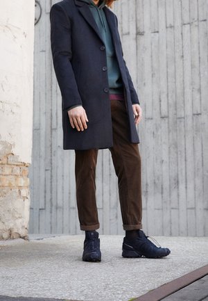 Personne portant un long manteau bleu marine, un sweat à capuche vert, un pantalon marron et des baskets noires, se tenant contre un mur en béton à l'extérieur.