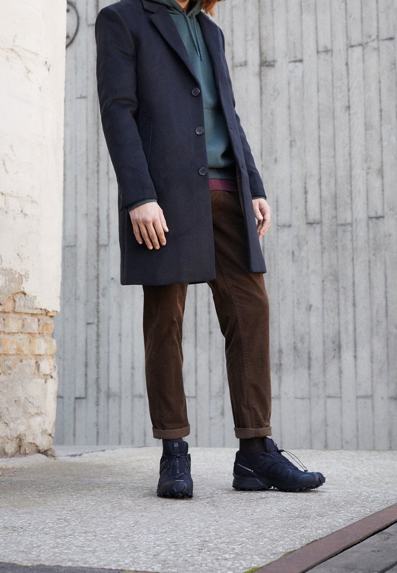 Person wearing dark navy overcoat, green hoodie, brown pants, and black sneakers standing against a concrete wall outdoors.