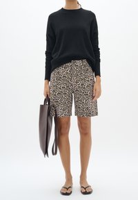 Femme portant un haut noir à manches longues, un short à imprimé léopard, des tongs noires et tenant un grand sac cabas marron foncé.