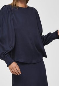 Mörkblå, långärmad blus i mjukt material. Har en rund halsringning och veckade puffärmar, med en avslappnad passform vid midjan.