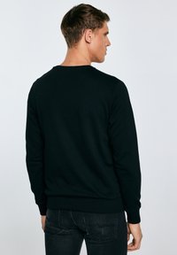 Mann trägt ein schwarzes Langarmshirt und dunkle Jeans, steht seitlich mit dem Kopf nach rechts gedreht vor einem schlichten Hintergrund.