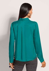 Vrouw met lang zwart haar, die grote oorbellen draagt, een teal lange mouwen blouse en zwarte gebloemde broek, met haar rug naar de camera gekeerd tegen een effen achtergrond.