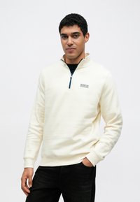 Krém színű pulóver, puha anyagból készült, félcipzáros nyakkal és tengerészkék logófolttal. Bordás mandzsetták és szegély.