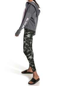 Šedá mikina s kapucí, na rukávu s nápisem "PUMA" v růžové barvě, kombinovaná s černými legínami s abstraktním bílým vzorem; černé sandály.