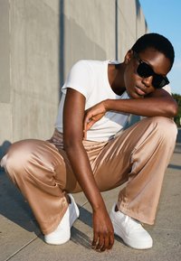 Junge Person mit schwarzen Sonnenbrillen, weißem T-Shirt, beigen Satin-Hosen und weißen Turnschuhen, die im Freien vor einer Betonwand hockt.