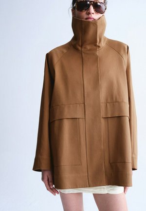 Persona que lleva un abrigo marrón extragrande con cuello alto y grandes bolsillos, combinado con gafas de sol carey y falda blanca.