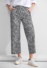 Osoba stojící s rukama v kapsách, má na sobě černobílé kalhoty s geometrickým vzorem trojúhelníků, bílou košili a bílé tenisky.