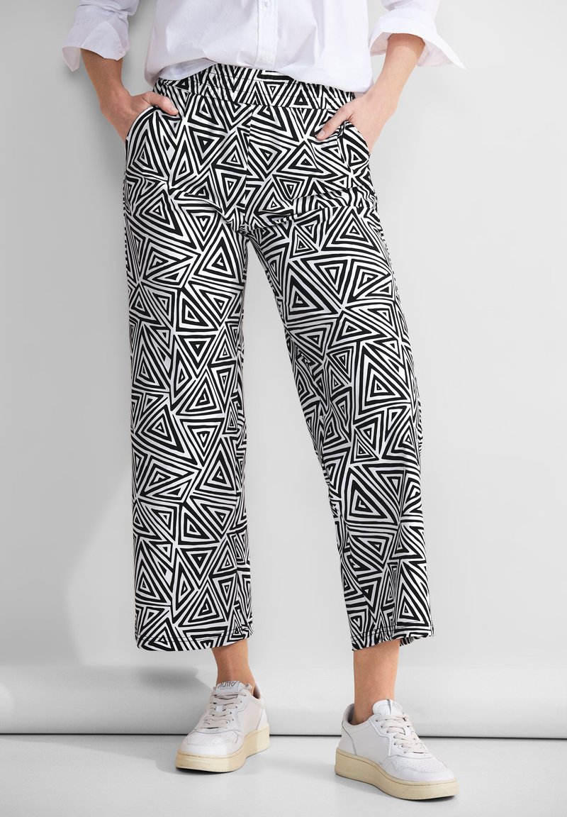 Osoba stojící s rukama v kapsách, má na sobě černobílé kalhoty s geometrickým vzorem trojúhelníků, bílou košili a bílé tenisky.