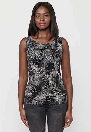 Haut sans manches noir avec un col drapé, orné d'un motif floral blanc et d'un design ajusté. Porté avec un jean foncé.