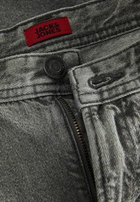 Närbild av grå denimjeans som visar en oöppen gylf, en knapp med präglad text, knapphål och en röd tygskylt med märkesnamn.