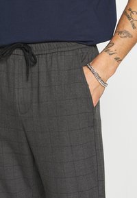 Man in een grijs geblokte broek met elastische tailleband en zwarte trekkoord, hand in zijn zak, getatoeëerde onderarm en een zilveren ketting armband.