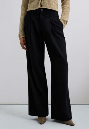 Los pantalones negros de pierna ancha presentan una textura suave y bolsillos laterales, combinados con un cárdigan beige texturizado. El conjunto se completa con unos tacones beige de punta.
