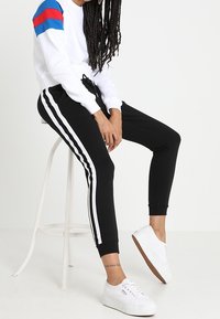 Vitt sweatshirts med blå och röda axelränder, kombinerat med svarta joggers med vita sidränder. Vita sneakers fulländar outfiten.