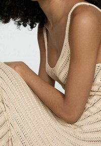 Donna con capelli ricci che indossa un vestito di maglia a uncinetto beige, seduta con le braccia appoggiate sulle ginocchia piegate su uno sfondo semplice.