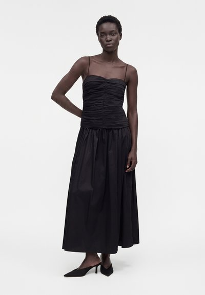 Γυναίκα που φοράει μαύρο τοπ με ρυτίδες και λεπτές τιράντες, μακριά μαύρη φούστα και μαύρα μυτερά Mule με τακούνι σε λευκό φόντο.