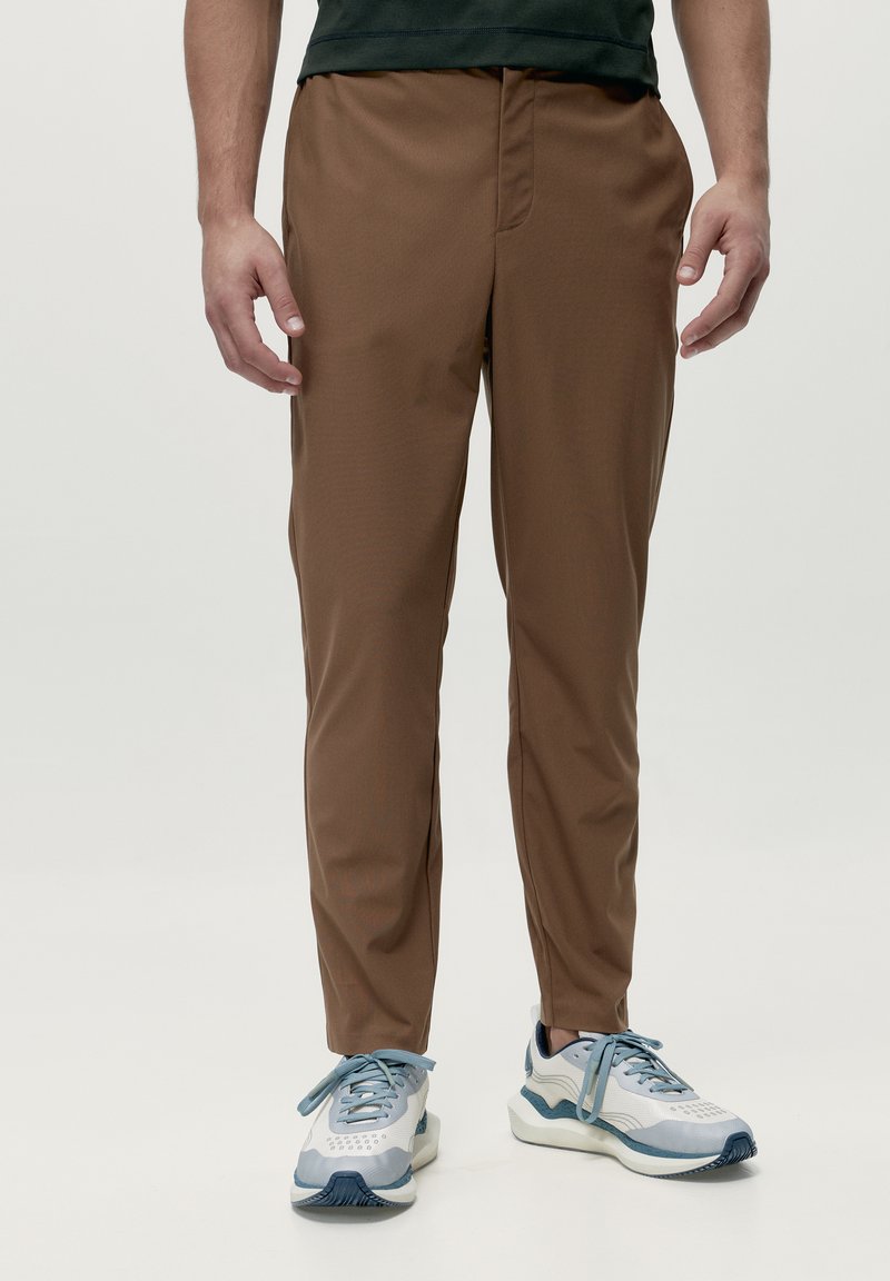 Brūnas, vieglas, sašaurinātas bikses ar gludu tekstūru un priekšējo rāvējslēdzēju. Saskaņotas ar gaiši ziliem sporta apaviem ar tumšiem akcentiem.