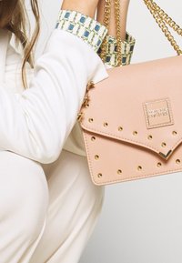 Žena v bílém obleku drží světle růžovou kabelku Versace Jeans Couture s kovovými řetízkami a vzorovanými manžetami košile.