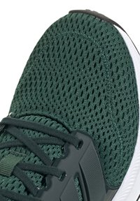 Zapatilla deportiva de malla verde con una superficie texturizada, detalles en negro y una suela de goma blanca. Incluye cordones negros y una lengüeta acolchada.